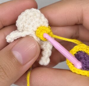 A cream crocheted flower held between finger and thumb, with yellow yarn wound round a pink needle at its centre