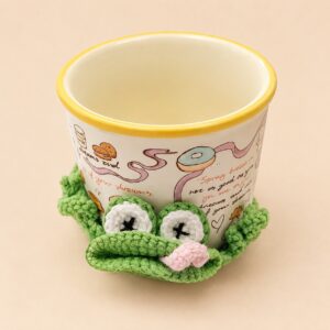 A crocheted bright green frog coaster under a cream mug with a yellow rim, the frog's head folded in half at the front edge with a pink tongue poking out of the fold, two white eyes ringed in green with a black cross stitched on each, and bobbly green legs curling up against the base of the mug on either side, on a plain peach background