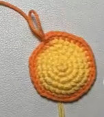 A flat crocheted circle of yellow single crochet with an orange edge, an orange yarn tail looped up from it, lying on a grey surface