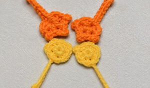 Two orange crocheted feet, each a fan of pointed toes at the end of a chain leg, above two small yellow crocheted wings with their yarn tails hanging down, on a grey surface