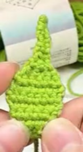 A flat green crocheted diamond with a narrow neck rising from its top, held up in the fingers in front of a labelled ball of green yarn
