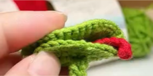 A green crocheted circle folded in half between finger and thumb, a red crocheted tongue tucked into the fold and curling out at one end