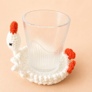 A crocheted white goose coaster with a ribbed drinking glass standing on it, the goose's head and neck rising from the left edge with an orange beak and a small black cross-stitched eye, and two orange webbed feet at the right