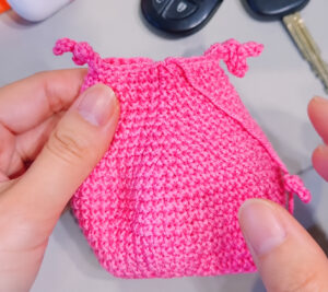 A pink crocheted fish pouch held in two hands before its lip and eyes go on: plain rounds, the tail end sewn closed with a little crocheted foot at each corner and a third foot at the side, with a car key fob and a key on the table behind
