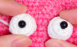 Two white crocheted eyes with black safety eyes, held against a pink crocheted pouch with the fingertips to find where they go