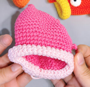 A pink crocheted fish pouch with a pale pink lip rolled back into a thick rim round its open mouth, held open in two hands, a finished orange fish with a yellow lip behind it