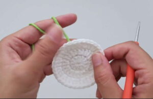 Two hands holding a small white crocheted circle in the middle of being made, a strand of green yarn already hooked over one finger and an orange-handled crochet hook resting against the other hand