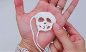 A small white crocheted skull with two eye sockets and a row of teeth lying in an open palm, its two long sewing tails hanging down and a sewing needle held between the fingers