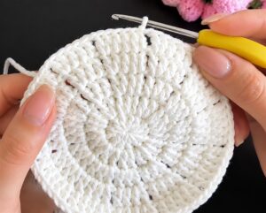 The white openwork centre of a crocheted coaster at the end of its fifth round, lines of small gaps running out from the middle, held in two hands with a yellow-handled hook in the work