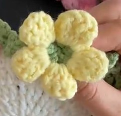A yellow crocheted flower with rounded petals sitting on the green edge of a white coaster, held between finger and thumb
