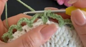 Green chain loops being crocheted round the edge of a white coaster centre, a yellow-handled hook in the work