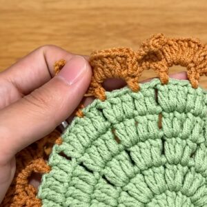 A thumb holding open the edge of a pale green crocheted body, with an orange edging of flat scallops and pointed leaves worked onto it