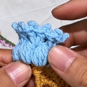 A finished pale blue crocheted lily held between two thumbs, its clusters gathered into a small bell with picots round the rim
