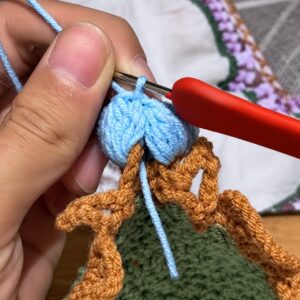 A crochet hook with a red handle working a pale blue lily's clusters into an orange scallop at the edge of a dark green crocheted piece