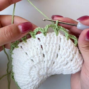 Green chain loops being crocheted round the edge of a white openwork coaster centre, the hook still in the work