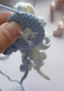 The blue base of a crocheted jellyfish held up in a hand, the white tails of the tentacles knotted through its centre and the tentacles hanging below
