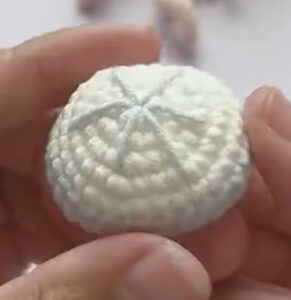 The white top of a crocheted jellyfish held between finger and thumb, pale lines stitched out from the centre of the dome
