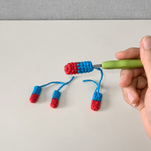 Four small crocheted limbs, red at the tip and blue above: three lying on a white table with long blue tails, the fourth still on a green-handled hook