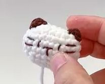 The cat's white crocheted head held in the fingers before it is sewn on: two small chestnut ears, and the eyes, nose and whiskers stitched in chestnut, with a white yarn tail hanging below