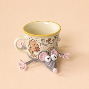 A cup standing on a crocheted mouse coaster in a white tray, the mouse's grey-brown head with a pink nose, pink-lined ears and cream eyes with black crosses poking out at the front, and four thin legs with pink paws spread out on either side