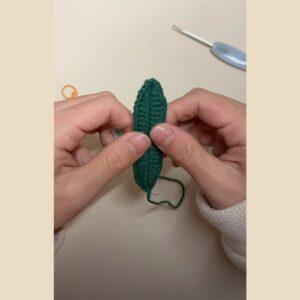 A finished dark green crocheted leaf held between two hands, its yarn tail hanging below, with a crochet hook and a stitch marker on the table behind