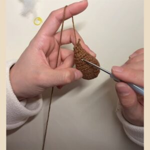 A small brown crocheted cone hanging from its yarn over a finger, the tip of a crochet hook going in under one of the rows round its outside
