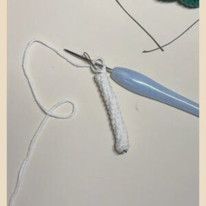 A narrow white crocheted tube still on the hook, its long yarn tail curling across the table, with a length of dark wire lying beside it