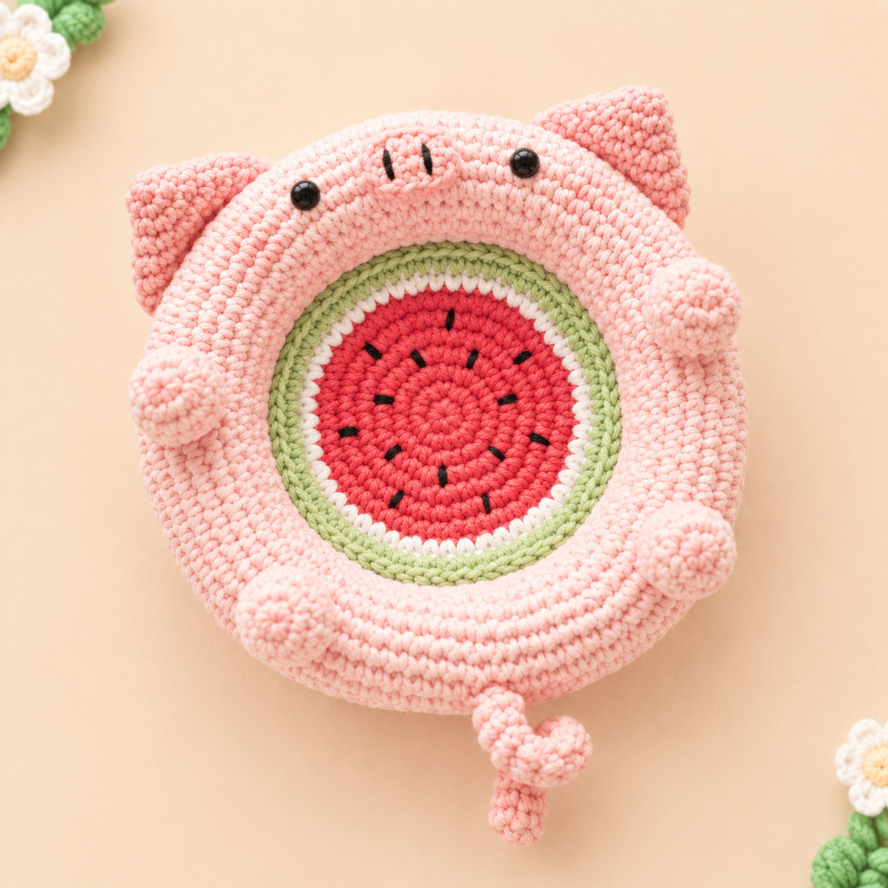 A pink crocheted pig dish seen from above on a peach background, with a watermelon slice in the well: red with short black seeds, a white ring and a green edge. The pig has two pointed ears, a snout with two black nostrils, black eyes, four round paws on the padded rim and a curly tail