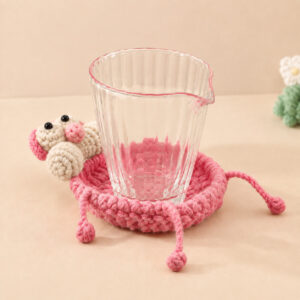 A crocheted pink panther coaster with a clear ribbed glass pitcher standing on it: a cream muzzle with a small pink nose and two cream eyes under pink eyelids with black pupils peek over one side of the rim, and thin pink arms and legs ending in bobbles dangle over the edge onto the table