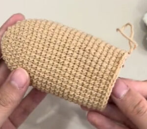 The tan body of a crocheted key pouch held in two hands before anything is sewn on: plain rounds, closed and rounded at one end, open at the other with the yarn still attached