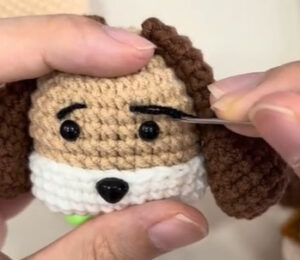 Black eyebrows being stitched onto a crocheted puppy's head with a needle, the black eyes, black nose, white muzzle and dark brown ears already on