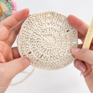 A crocheted coaster centre in cream, held up in two hands at the end of its fifth round, small holes lined up in spokes, a pale yellow hook in one hand