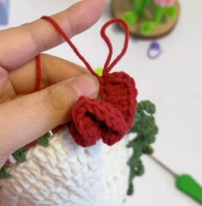 A red strip of double crochets standing up from the leaf edging of a white coaster centre, held between finger and thumb before it is rolled into a rose