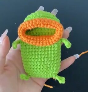 A green crocheted pouch held in a palm with its orange lip picked up round the mouth and rolled outwards into a thick rim, its little arms and legs tied on