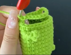 A green crocheted pouch held on its side in the fingers, still open at the top, with a row of chains arching across one edge to leave the mouth opening and a red-handled hook still in the work