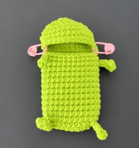 A lime-green crocheted pouch laid flat before its lip goes on: the top closed, two little legs and two arms tied on, and the mouth opening across the upper front with a pink stitch marker at each corner