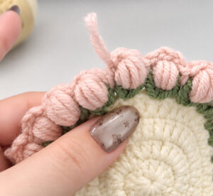 Pink puff stitches sitting in the green V's round the edge of a cream crocheted disc, each a plump bundle of long loops, with a pink yarn tail standing up from one of them