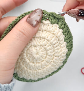 A green round of V's being crocheted round the edge of a cream disc, held in the fingers with a grey crochet hook in the work at the top