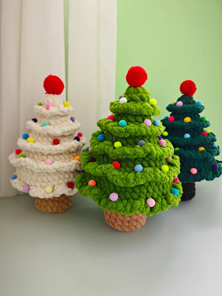 Three crocheted chenille Christmas trees standing in front of a white curtain and a pale green wall: a cream one, a bright green one in front and a dark green one behind, each with tiers of ruffled frills, small pompoms in mixed colours, a big red pompom on top and a short crocheted trunk