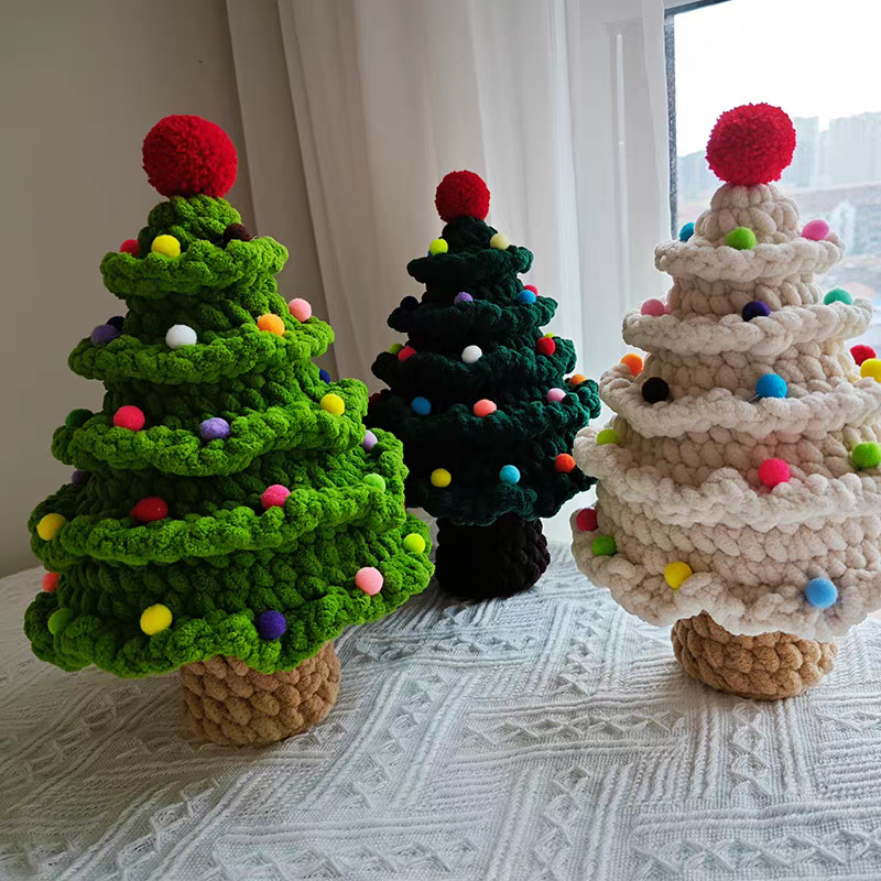 Three crocheted chenille Christmas trees on a white textured blanket beside a window with white curtains: bright green on the left, dark green behind and cream on the right, each with tiers of ruffled frills, small pompoms in mixed colours and a big red pompom on top