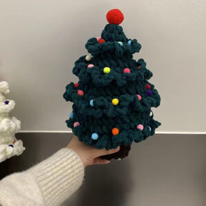 A crocheted Christmas tree in dark green chenille held up on one hand in front of a white wall: four tiers of ruffled frills dotted with small pompoms in pink, yellow, blue, orange and white, a big red pompom on top, and a short dark brown trunk