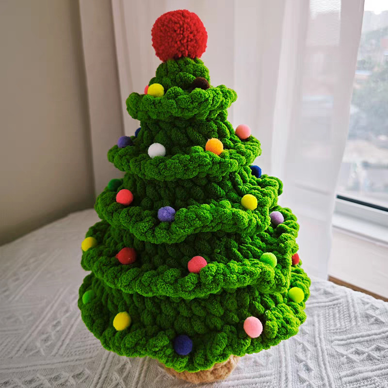 A crocheted Christmas tree in bright green chenille on a white textured blanket beside a window with white curtains, its tiers of ruffled frills dotted with small pompoms in yellow, pink, red, purple, orange, white and blue, with a big red pompom on top