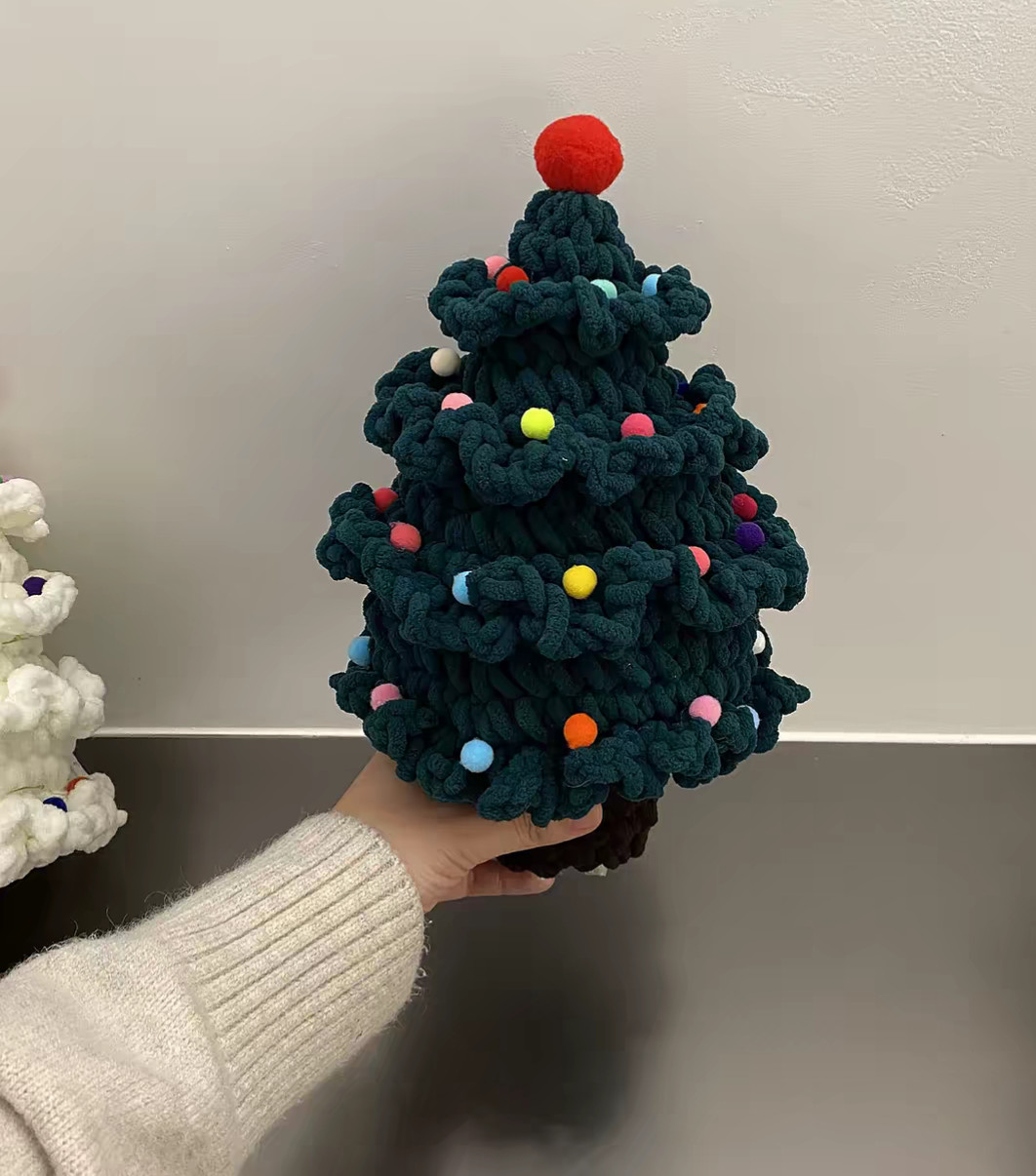 A crocheted Christmas tree in dark green chenille held up on one hand in front of a white wall: four tiers of ruffled frills dotted with small pompoms in pink, yellow, blue, orange and white, a big red pompom on top, and a short dark brown trunk