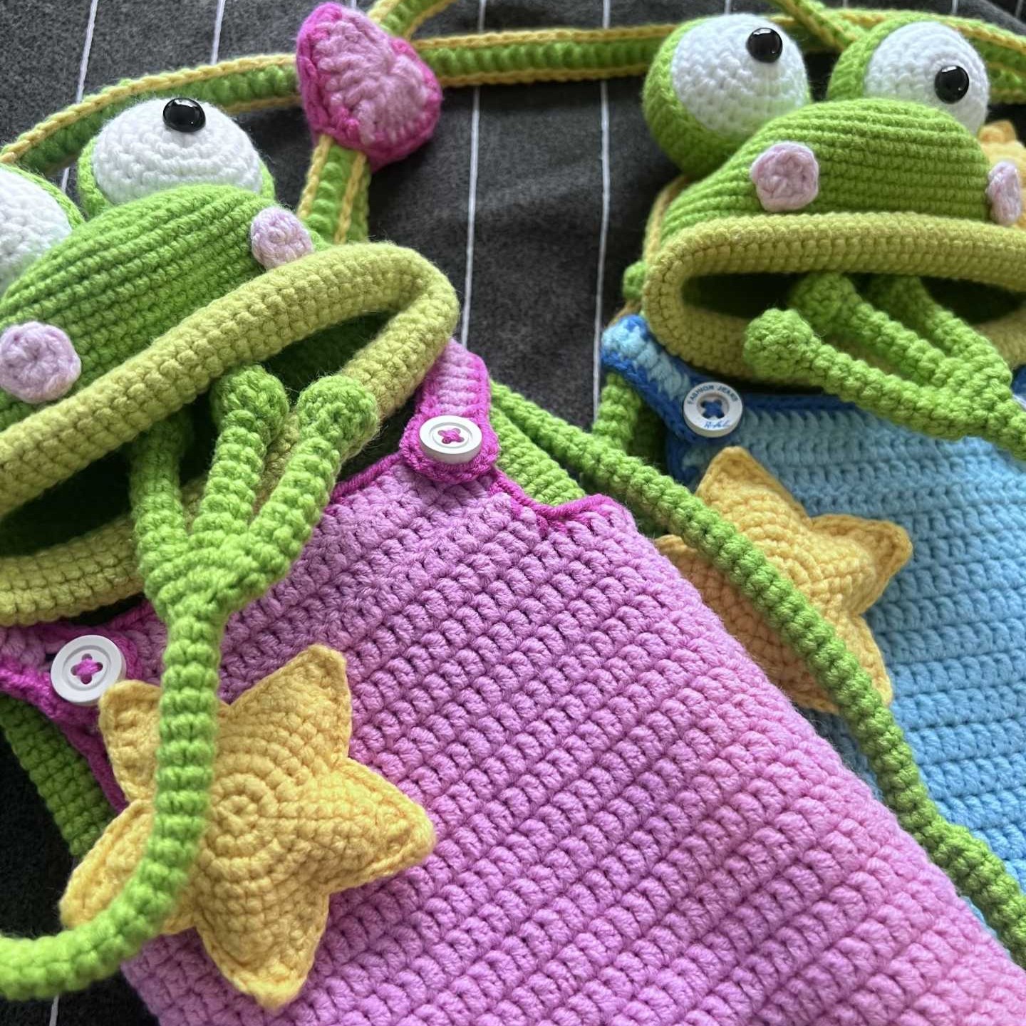 A close-up of two crocheted green frog bags on dark striped fabric: the nearer one in bright pink dungarees with a yellow star and two white buttons, its long green arms trailing out of its wide open mouth; the other in blue dungarees with a yellow star; both with white goggle eyes with black pupils and lilac cheeks