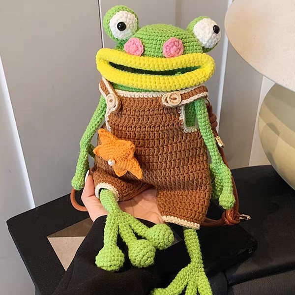 A crocheted green frog bag held up on one hand, in brown dungarees edged in cream with an orange star on the front, a bright yellow lip round its wide mouth, pink cheeks and white goggle eyes; its long green legs with three round toes hang over the hand, and a brown leather strap is clipped on behind