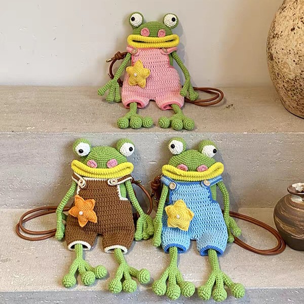 Three crocheted green frog bags sitting on stone steps, each with goggle eyes, pink cheeks, a wide yellow mouth, a star on its front and long arms and legs: one in pink dungarees on the upper step, and one in brown and one in blue on the step below, with brown leather straps coiled beside them