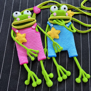 Two crocheted green frog bags lying side by side on black pinstriped fabric, one in pink dungarees and one in bright blue, each with white goggle eyes, pink cheeks, a wide open mouth with an olive green lip, a yellow star on its front, white buttons on its straps, and long thin arms and legs ending in round toes; green cord straps edged in yellow loop above them, with a pink crocheted heart on one
