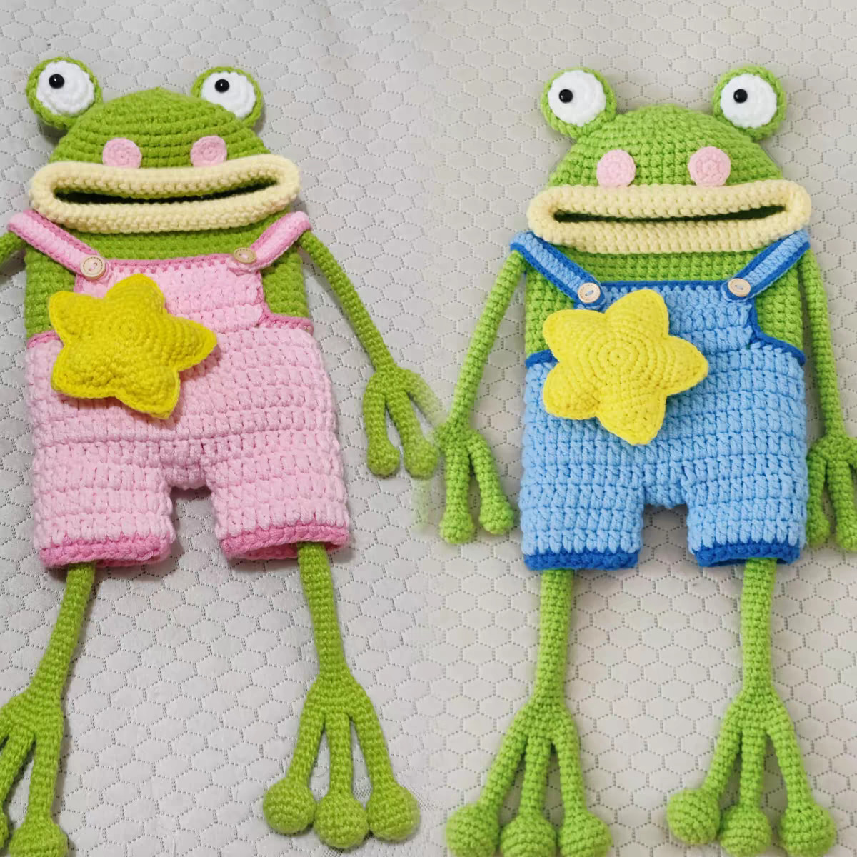 Two crocheted green frog bags laid flat on a white quilted surface, one in pale pink dungarees and one in light blue dungarees edged in darker blue, each with white goggle eyes, pink cheeks, a wide pale yellow lip round its mouth, a yellow star on the front, two wooden buttons, long thin arms ending in three fingers and long legs ending in three round toes