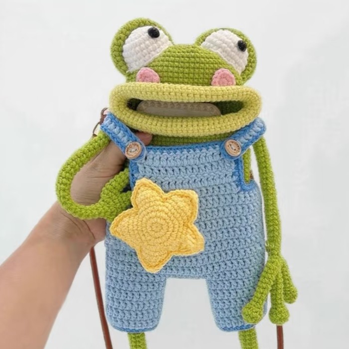 A crocheted green frog bag held up in one hand against a white background, in light blue dungarees edged in darker blue with a yellow star on the front and two wooden buttons, the top of a phone showing inside its wide open mouth; it has white goggle eyes, pink cheeks and long green arms, and a brown strap hangs down behind