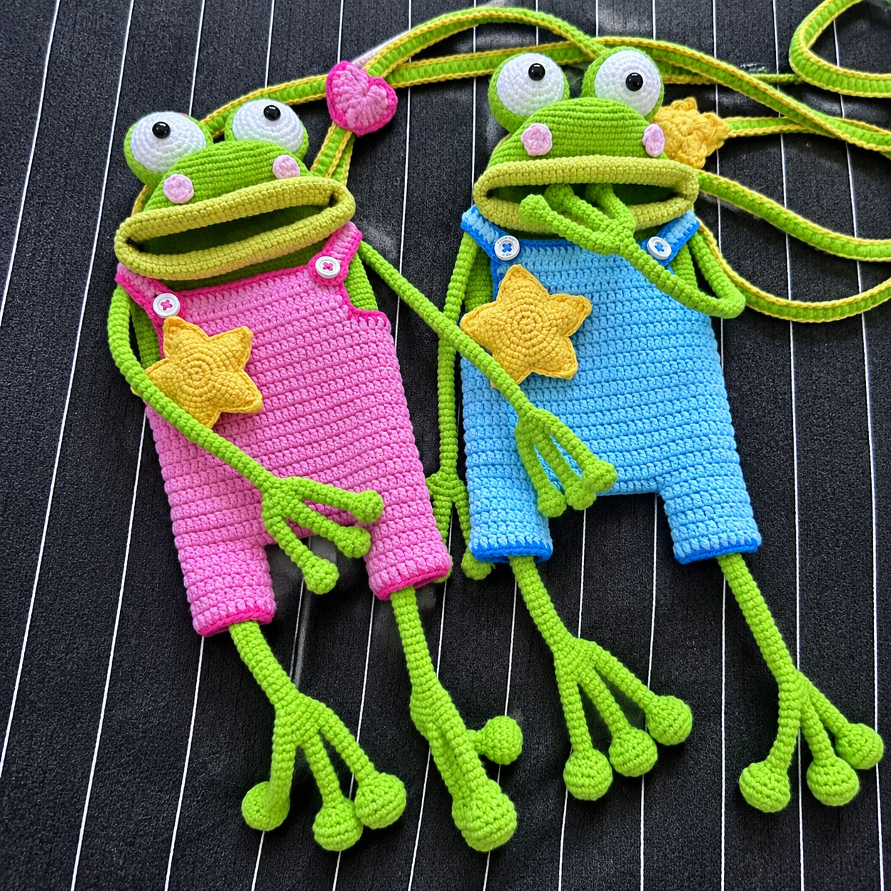 Two crocheted green frog bags lying side by side on black pinstriped fabric, one in pink dungarees and one in bright blue, each with white goggle eyes, pink cheeks, a wide open mouth with an olive green lip, a yellow star on its front, white buttons on its straps, and long thin arms and legs ending in round toes; green cord straps edged in yellow loop above them, with a pink crocheted heart on one
